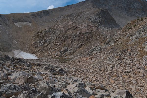 Half an hour later, I encounter the upper of the two tarns alternating my path between the ridge and the drainage.  Once again, I move right (N) to re-gain the ridge, avoiding boulder-hopping.  The saddle is above and left (E) of the highest snow in the couloir.  The route crosses under the buttress (in shadow) in the upper right of the frame.  The route unfolds as one progresses, working its way up through the broken rock buttress, well above the tarn, toward the higher snowfield.
