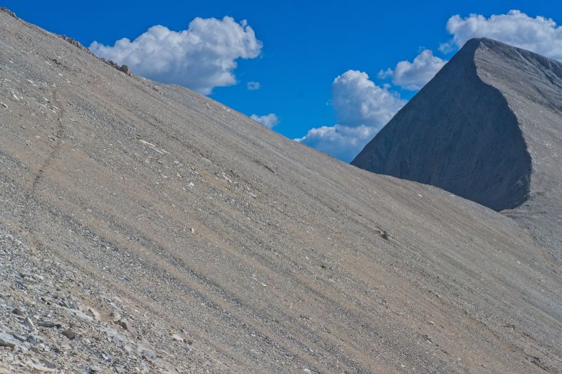 The game trail leading from the outcrop to the upper saddle is clearly evident on the left side (north) of the frame, intersecting the saddle at a point under the cloud.
