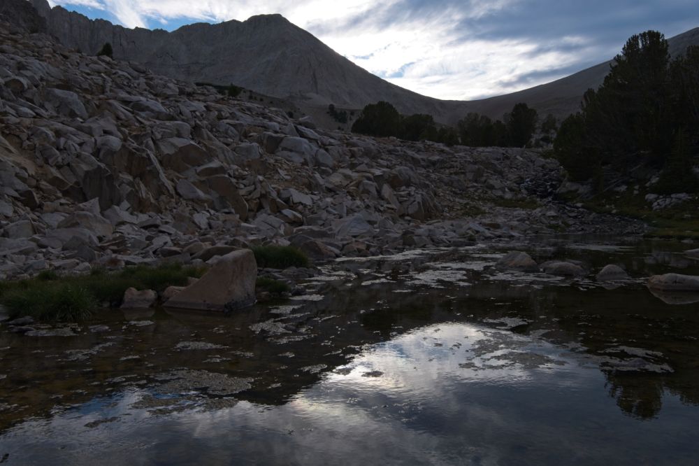 The saddle between D. O. Lee Peak and WCP-9, backlit, but dramatic high clouds have improved the sky, and their reflections are seen in the inlet stream adding interest in the foreground.
